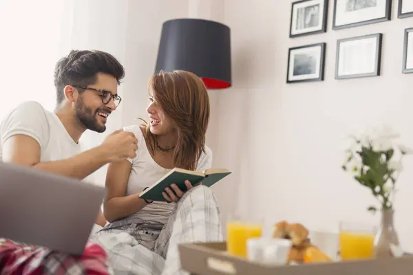 stock image Beautiful young couple in love having fun reading and surfing the net while having breakfast in bed. Focus on the girl
