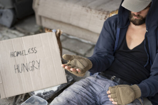 Homeless man wearing hoodie over cap and fingerless gloves, sitting on the street, begging for money. Focus on the hand
