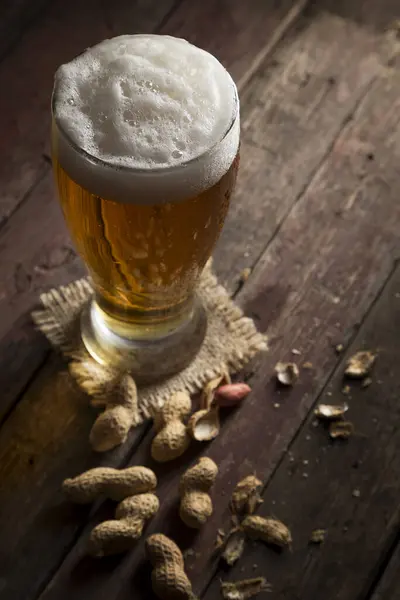 バーラップコースターの上に置かれた冷たい淡いビールのガラスと素朴な木製のテーブルにピーナッツ 泡に焦点を当てる — ストック写真