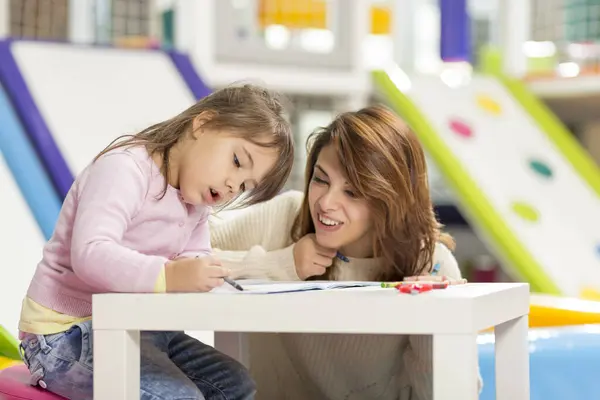 Mother and daughter sitting on a playroom floor, drawing and coloring with creons. Focus on the daughter