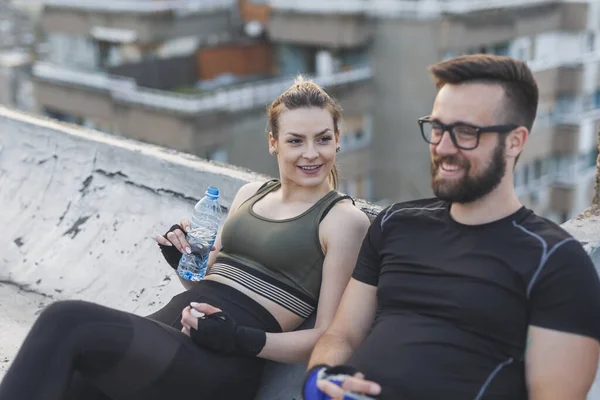 Couple sitting on a building rooftop terrace, taking a break from a hard workout, having a conversation and drinking water