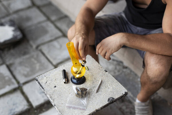 Молодой человек, курящий травку с помощью бонга; подробности мужской стороны заполнения бонг с марихуаной, получая его готов к использованию. Выборочный фокус на пальцах и горшке в кальяне