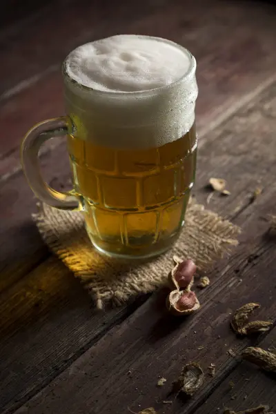 Mug of light beer with foam placed on a burlap cover and some peanuts on a rustic wooden pub table. Selective focus on the foam