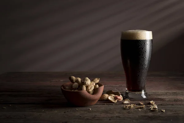 一杯冷而黑的啤酒 泡沫放在一张乡村木桌上 旁边放着一碗花生 — 图库照片