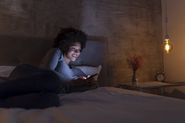 Güzel, genç, melez bir kadın yatakta uzanıyor ve akıllı telefondan mesaj yazıyor.