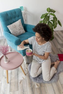 Güzel, melez bir kadının oturma odasında oturup kahvaltılık gevreğe süt dökerek evde boş vaktinin tadını çıkardığı yüksek açılı bir manzara.