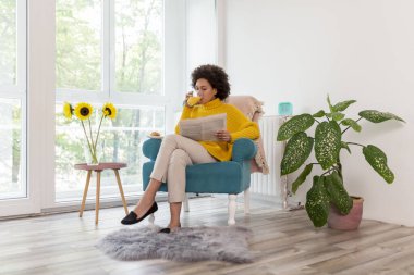 Güzel, genç, melez bir kadın evde dinleniyor, bir koltukta oturuyor, gazete okuyor ve kahvaltı yapıyor, taze sıkılmış portakal suyu içiyor.