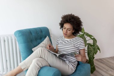 Güzel, genç, melez bir kadın koltukta oturuyor, dergi okuyor ve evde dinleniyor.