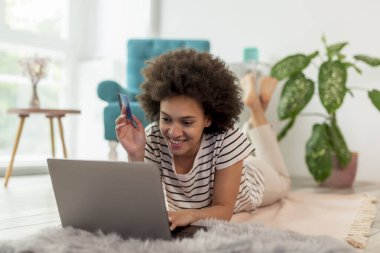 Güzel, genç, melez bir kadın oturma odasında yerde yatıyor internetten para harcıyor, bilgisayarla internet alışverişi yapıyor, kredi kartıyla ödüyor.
