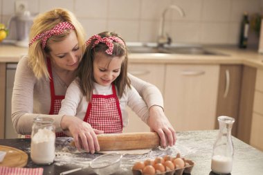 Anne ve kızı mutfakta kurabiye pişiriyor, küçük kız merdane kullanıyor, annesinin yardımıyla kurabiye hamurunu yoğuruyor.
