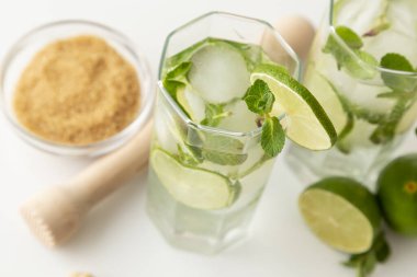 İki mojito kokteyli, bol buzlu, beyaz rom, limon suyu ve tonik, modern beyaz bir masa üzerinde limon dilimleri ve nane yapraklarıyla süslenmiş. Bardaktaki nane yapraklarına odaklan.