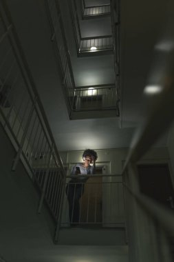 Genç bir kadın apartman koridorunda merdivenlerde durup müzik dinliyor, kulaklık takmış ve akıllı telefon listesinden bir şarkı seçiyor.