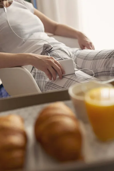 Detail of a woman wearing pajamas, sitting in bedroom, holding a mobile phone and listening to the music. Breakfast tray in the foreground. Focus on the phone and the hand