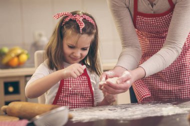 Mutfak masasına un serpiştiren küçük bir kız annesine pizza hamuru pişirmesinde yardım ediyor.