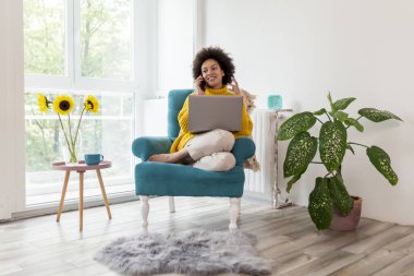 Güzel melez bir kadın koltukta oturuyor, evinde dinleniyor, dizüstü bilgisayar kullanıyor ve telefonla konuşuyor.