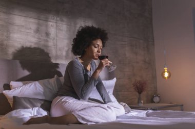 Pijamalı bir kadın yatakta oturmuş, şarap içiyor ve evde boş vakitlerin tadını çıkarıyor.