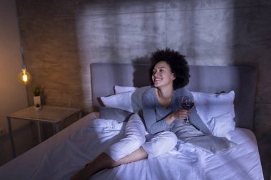 Genç melez kadın pijama giyiyor, bir bardak şarap içiyor ve yoğun bir günün ardından evde dinleniyor.