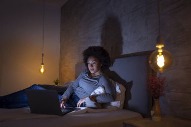 Genç ve güzel melez bir kadın geceleri yatakta dizüstü bilgisayarla internette sörf yapıyor.