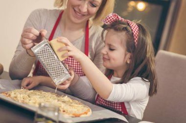 Anne ve kızı mutfakta pizza yapıyor; kızı fırında pişmeden önce peyniri rendeliyor.