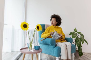 Güzel, melez, genç bir kadın bir sandalyede oturuyor, atkı örüyor ve evde dinleniyor.