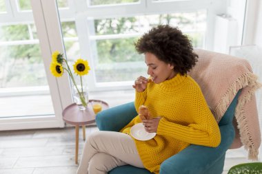 Güzel, genç, melez bir kadın evde dinleniyor, bir koltukta oturuyor ve kahvaltıda taze kruvasan yiyor.