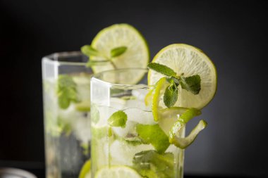İki mojito kokteyli, bol buzlu, beyaz rom, limon suyu ve tonik, limon dilimleri ve nane yapraklarıyla süslenmiş. Bardaktaki nane yapraklarına odaklan.