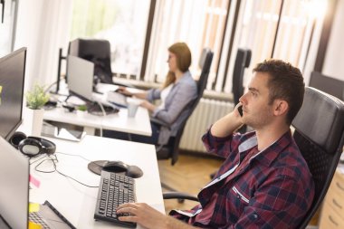 İki iş arkadaşı bir ofiste masalarında oturuyor, çalışıyor ve telefon görüşmeleri yapıyor; yazılım geliştiricileri kodlama yapıyor