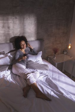 Pijamalı bir kadın yatağa uzanıyor, şarap içiyor ve evde boş vakitlerin tadını çıkarıyor.