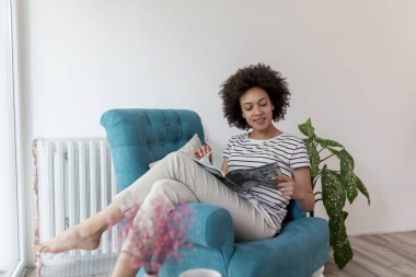 Güzel, genç, melez bir kadın pencerenin kenarında oturmuş dergi okuyor ve evde eğleniyor.