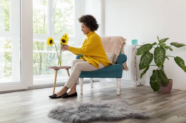 Güzel melez genç bir kadın oturma odasının penceresinin yanındaki koltukta oturuyor, vazoya ayçiçeği buketi yerleştiriyor, evde boş vakitlerin tadını çıkarıyor.