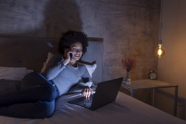Güzel, genç, melez bir kadın geceleri yatakta dizüstü bilgisayar kullanıyor ve telefonla konuşuyor.