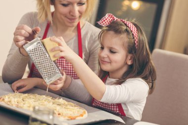 Anne ve kızı mutfakta pizza yapıyor; kızı pişirmeden önce üzerinde peynir rendeliyor. Kızına odaklan.