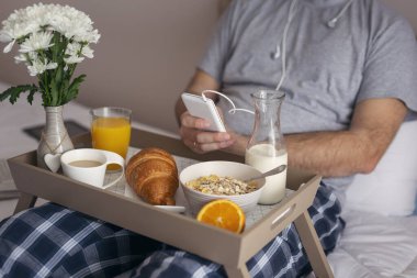 Erkek eli kulaklıkla akıllı bir telefon tutuyor yatakta kahvaltı yaparken müzik dinliyor. Telefona odaklan.