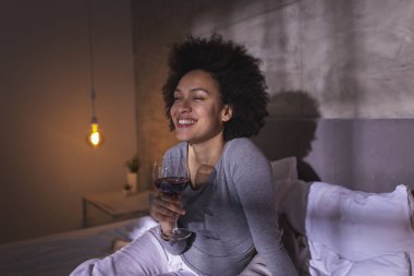 Pijamalı bir kadın yatakta oturmuş, şarap içiyor ve evde boş vakitlerin tadını çıkarıyor.