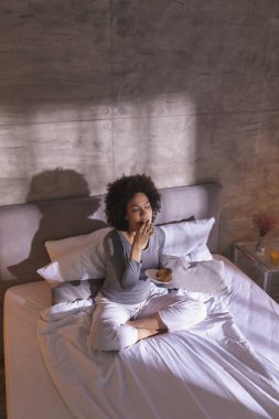 Yüksek açılı, genç, melez bir kadının pijama giyip, sabah kahvaltısını yapıp, evde dinlendiği bir manzara.