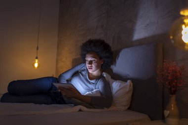 Güzel, genç, melez bir kadın geceleri yatakta yatıp tablet bilgisayarında internette sörf yapıyor.