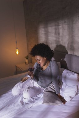 Güzel melez bir kadın pijamalarıyla yatakta oturuyor, bir bardak şarap içiyor ve evde dinleniyor.