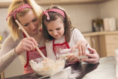 Güzel anne-kız mutfakta eğleniyor; anne hamur yoğururken kız çocuğu, mutfak kaşığıyla un ekliyor.