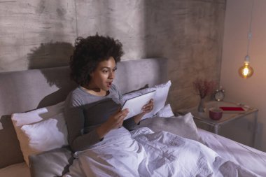 Pijama giyen, sabahları yatakta uzanan, tablet bilgisayar kullanan, haber aldıktan sonra şaşıran, güzel, melez genç bir kadın. 