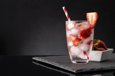 Bir bardak taze çilek dilimli soğuk su ve siyah taş tepside bir kase taze çilek.