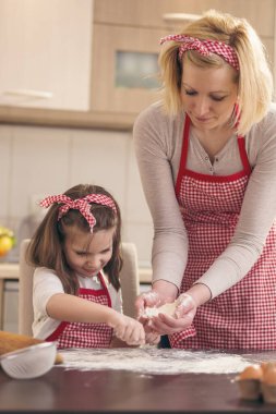 Mutfak masasına un serpiştiren küçük bir kız annesine pizza hamuru pişirmesinde yardım ediyor.