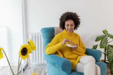 Güzel melez kadın evde dinleniyor, koltukta oturuyor ve kahvaltı yapıyor.