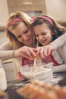 Genç anne mutfakta kızıyla hamur pişiriyor, bir yumurta kırıyor ve kız da unu karıştırıyor.