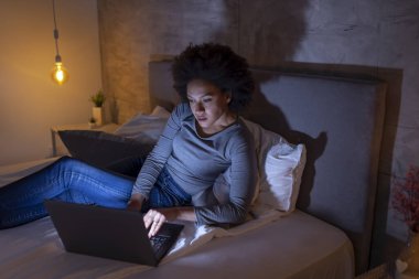 Güzel, genç, melez bir kadın geceleri yatağında dizüstü bilgisayar kullanarak geç saatlere kadar çalışıyor.