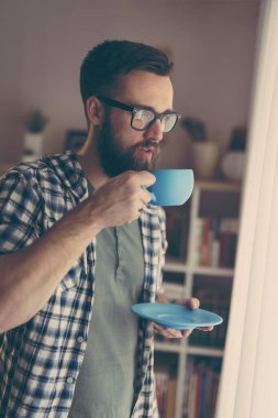 Pencerenin yanında duran yakışıklı adam, kahve içiyor.