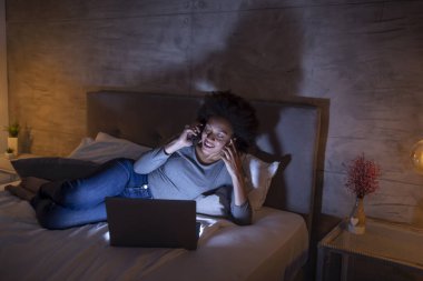 Güzel, genç, melez bir kadın geceleri yatakta dizüstü bilgisayar kullanıyor ve telefonla konuşuyor.