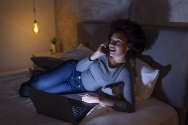 Güzel, genç, melez bir kadın geceleri yatakta dizüstü bilgisayar kullanıyor ve telefonla konuşuyor.