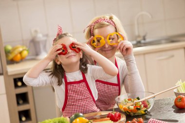 Güzel anne ve kız mutfakta salata yaparken, biber dilimlerini bardak gibi tutarken ve dikizlerken eğleniyorlar. Kızına odaklan.