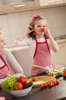 Anne ve kız sebze kesiyor, salata yapıyor ve eğleniyor; kız, küpe ve burun olarak vişneli domates yerleştiriyor.