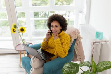 Güzel melez bir kadın koltukta oturuyor, evinde dinleniyor, dizüstü bilgisayar kullanıyor ve telefonla konuşuyor.